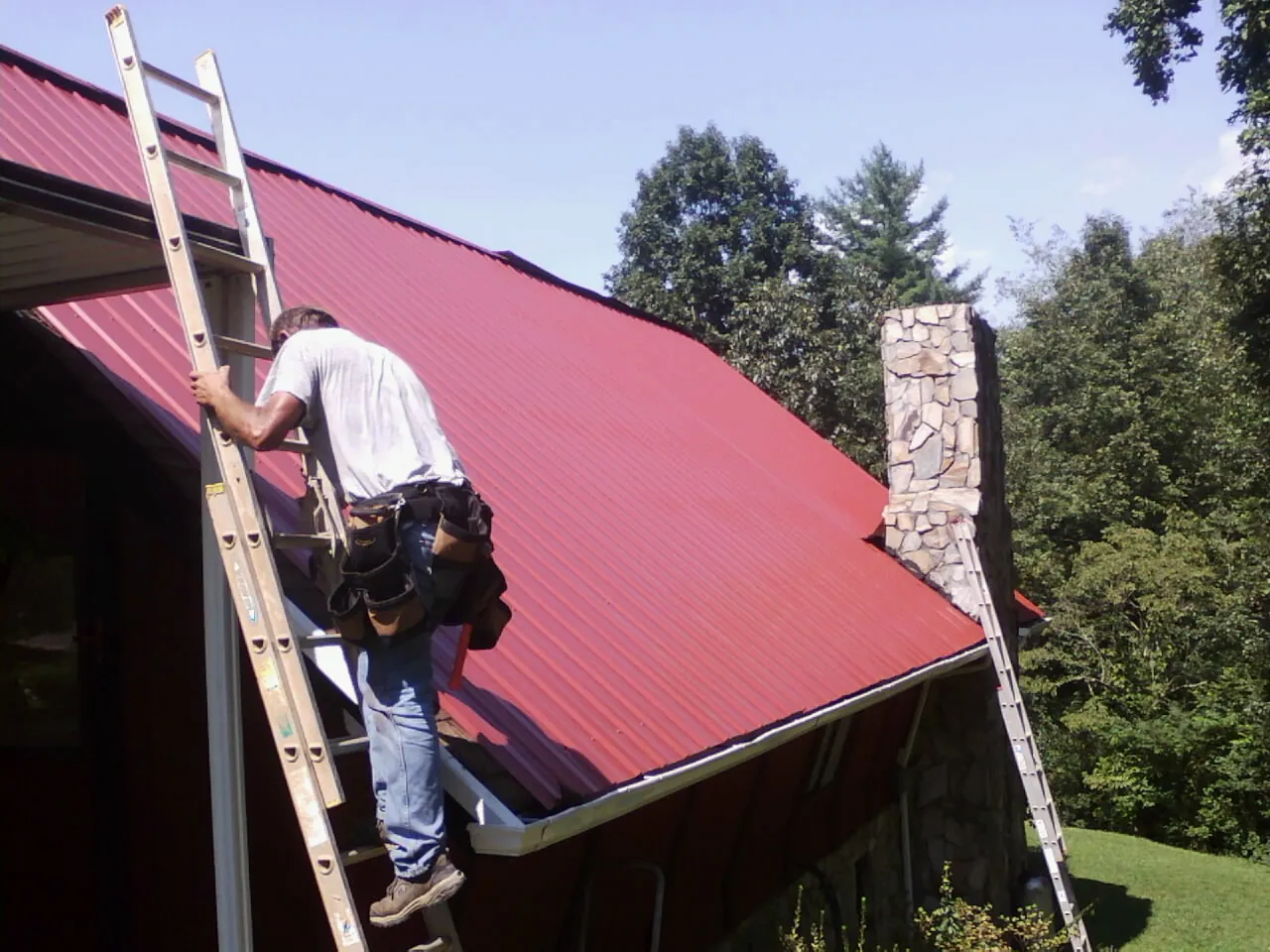 Roofer on a ladder beside a red metal roof and stone chimney — Soffit & Fascia Repair by Roofing Brothers in Laramie